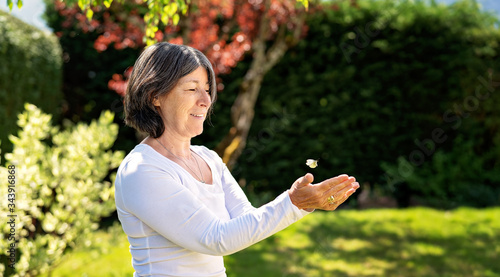 Happy senior woman releasing butterfly letting it fly from her hands in garden at bright summer day. Proximity to nature. Active senior lifestyle, happiness and support concept. Beauty of nature