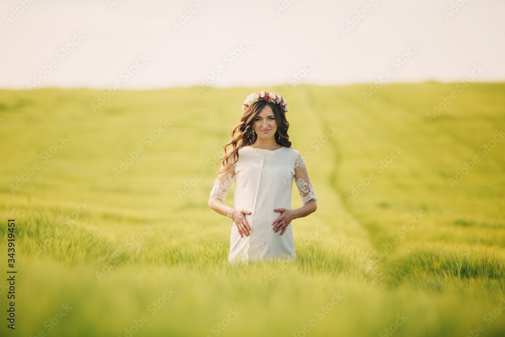 pregnant woman in white dress with wreath of flowers on head is keeping her hands on stomach and relaxing outdoors in grass field. motherhood concept, selective focus pregnant woman in white dress with wreath of flowers on head is keeping her hands on stomach and relaxing outdoors in grass field. motherhood concept, selective focus