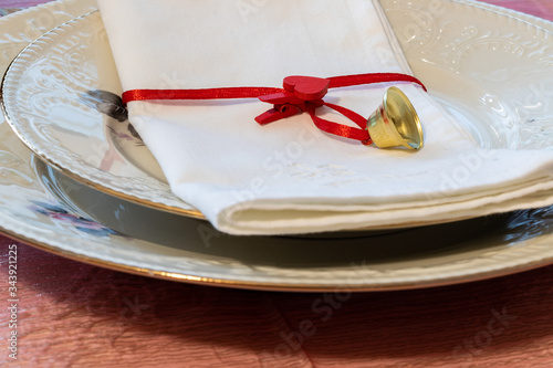 Preparations for the end of year dinner at home. A set table with a napkin closed by a small heart-shaped clothespin and a golden bell on two porcelain plates.