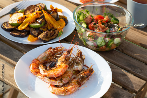 grilled langoustines and vegetables on white plates, a bowl with fresh salad on a garden wooden table outdoor. Summer time