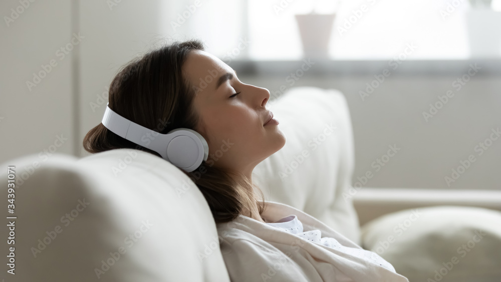 Peaceful girl in modern wireless headphones sit relax on comfortable couch  listening to music, happy calm young woman in earphones rest on cozy sofa,  enjoy good quality sound, stress free concept Stock