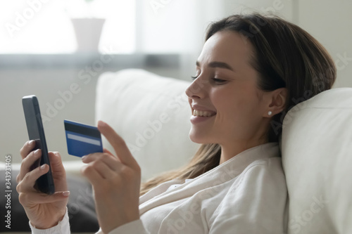 Smiling millennial girl relax on couch at home shopping online on smartphone using credit card, happy young woman make Internet order purchase on cellphone, enter banking service system on gadget