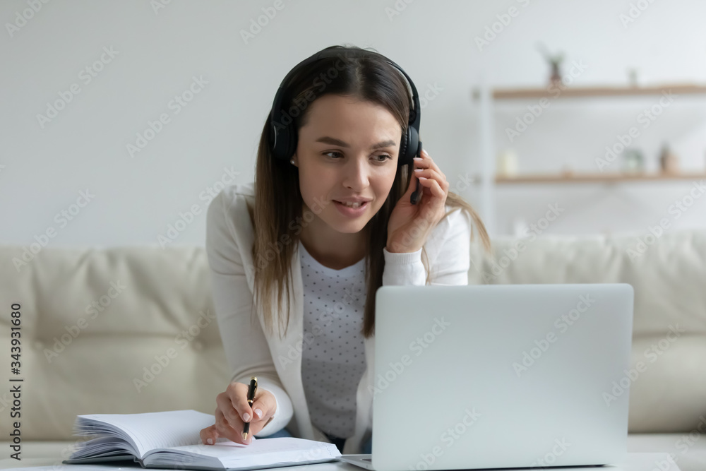 Young woman in earphones sit on couch at home study online on laptop, millennial girl in headphones take Internet course or training, make notes, watch webinar on computer, distant education concept