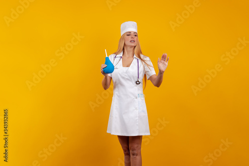 Emotional young woman doctor nurse in a white coat and hat with a douche enema in hands posing on a yellow background photo