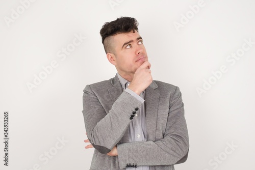 Portrait of thoughtful smiling man keeps hand under chin, looks sideways, thinking or wondering about something with interest, dressed casually, poses against gray studio. Taking decisions concept.