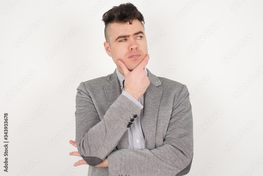 Isolated portrait of stylish young European man with hand under chin and looking sideways with doubtful and skeptical expression, worry and doubt. Standing indoors over gray background. Isolated portrait of stylish young European man with hand under chin and looking sideways with doubtful and skeptical expression, worry and doubt. Standing indoors over gray background.