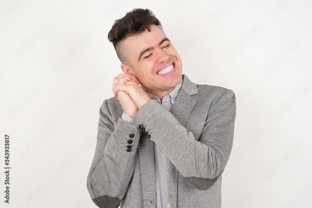 Dreamy young male with pleasant expression, closes eyes, keeps hands crossed near face, thinks about something pleasant, poses against gray background. Dreamy young male with pleasant expression, closes eyes, keeps hands crossed near face, thinks about something pleasant, poses against gray background.