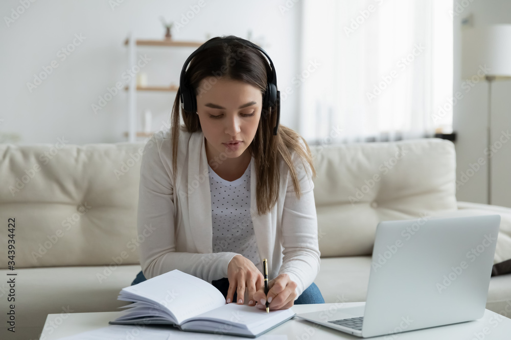 Concentrated millennial girl in headphones sit on couch at home make notes studying online on laptop, focused young woman watch webinar or Internet course on computer, distant education concept Concentrated millennial girl in headphones sit on couch at home make notes studying online on laptop, focused young woman watch webinar or Internet course on computer, distant education concept