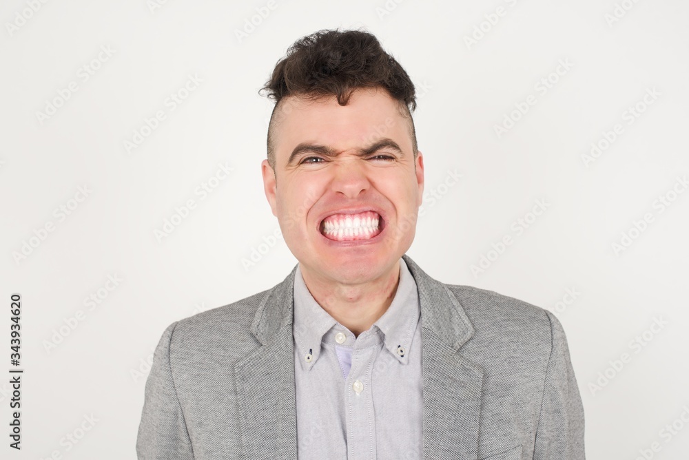 Mad crazy man clenches teeth angrily, being annoyed with coming noise, dressed in fashionable clothes, isolated over gray background. Negative feeling concept. Mad crazy man clenches teeth angrily, being annoyed with coming noise, dressed in fashionable clothes, isolated over gray background. Negative feeling concept.