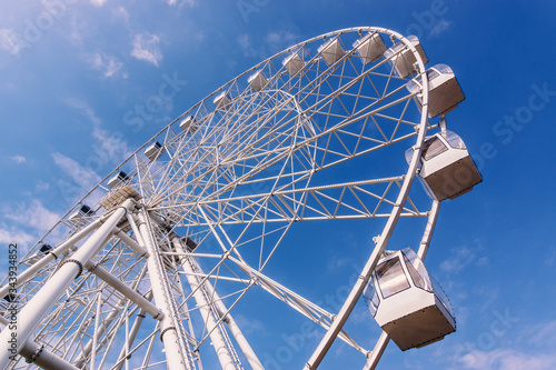 White metal construction of an attraction Ferris wheel with observation cabs high against a blue sky with white clouds.