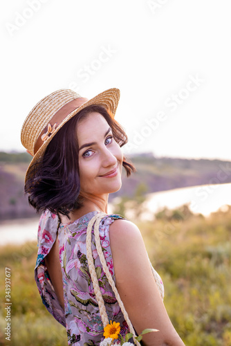 Portrait of romantic short haired smiling brunette woman in straw hat on a head with flowers in bag in amazing sunny field.Warm sunset colors.Wind play with hair.Concept of picnic date