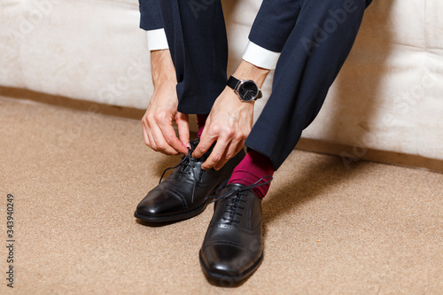 A businessman in dark blue suit, red socks and watches tying his shoelaces of black shoes. Modern, stylish and expensive look for young man