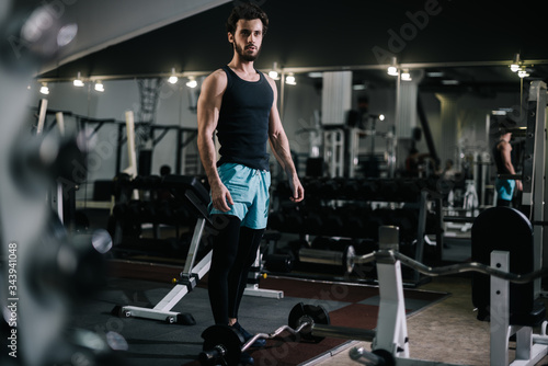 Athletic bearded young man with muscular wiry body wearing sportswear posing in modern dark gym. Concept of healthy lifestyle.