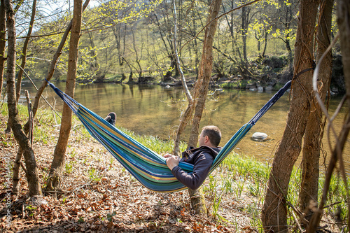 Young man is lying in a colorful hammock  near the trees  a river and mountainse early in the spring. He is alone and have a relaxing time in the nature