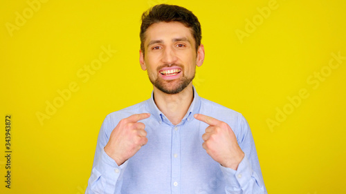 Choose me! Handsome bearded man shows his fingers on a yellow background with copyspace. Guy in a light blue shirt with his hands up. Place for text or product