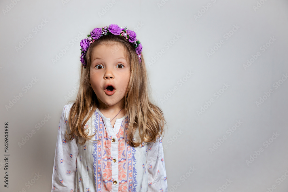 Portrait of pretty little girl in white dress and purple wreath on gray isolated background. Wonder and wow emotion concept Portrait of pretty little girl in white dress and purple wreath on gray isolated background. Wonder and wow emotion concept