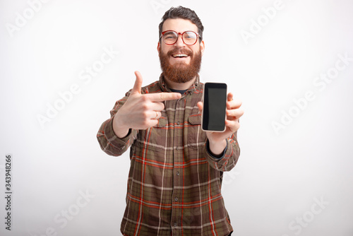 Smiling man wearing glasses and beard is pointing on his phone on white background.