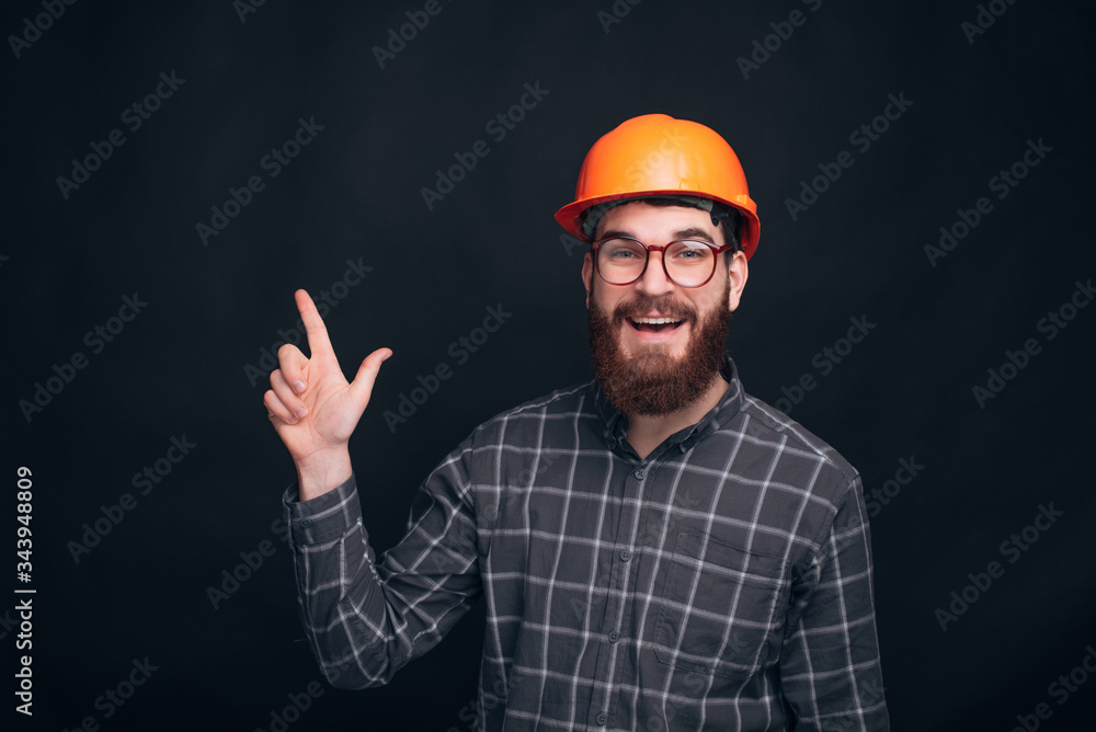 Smiling engineer or architect man wearing orange hard hat is pointing up on black background. Smiling engineer or architect man wearing orange hard hat is pointing up on black background.