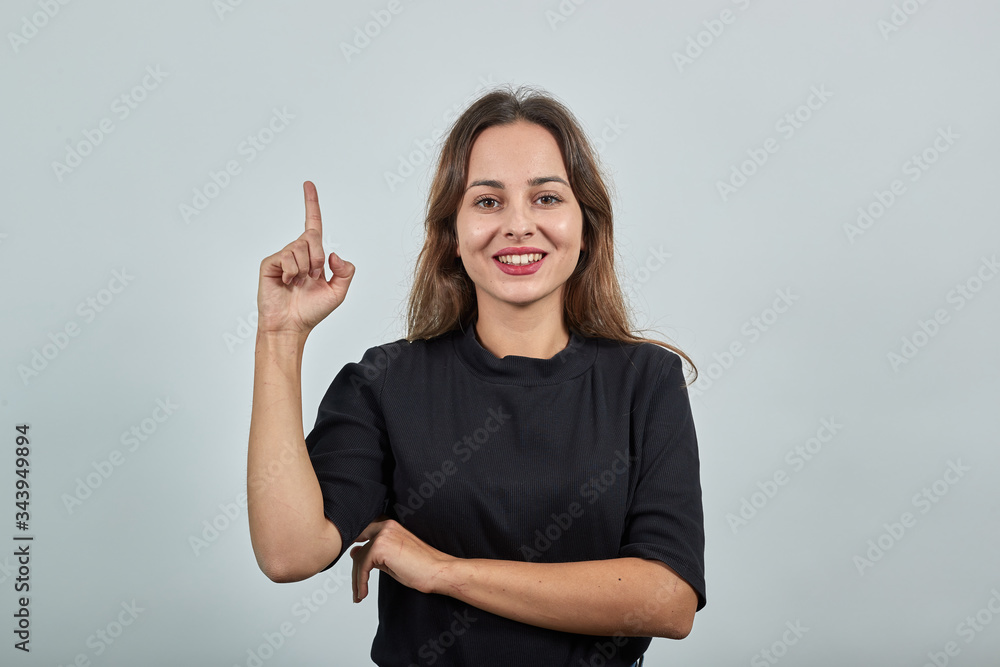 Cute young brunette woman in black t-shirt, blue jeans with belt on gray background, smiling happy girl raised her index finger up from a new idea Cute young brunette woman in black t-shirt, blue jeans with belt on gray background, smiling happy girl raised her index finger up from a new idea