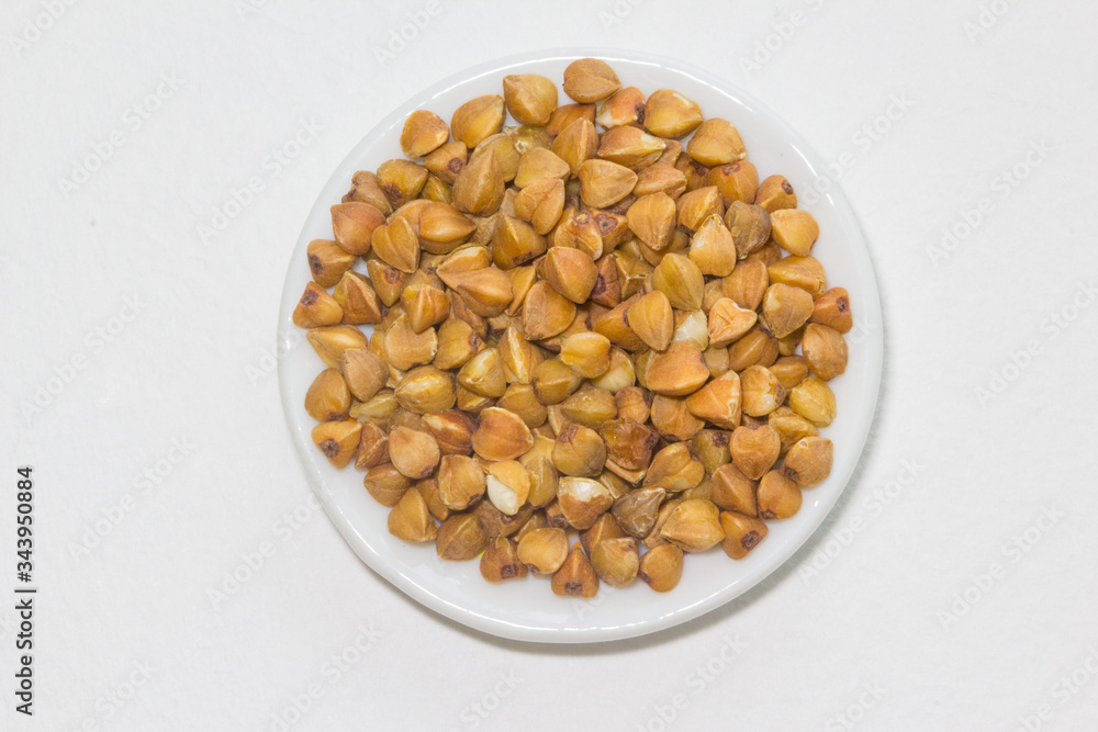 Raw buckwheats on the plate on the white background. Closeup plate with buckwheats. Raw buckwheats on the plate on the white background. Closeup plate with buckwheats.