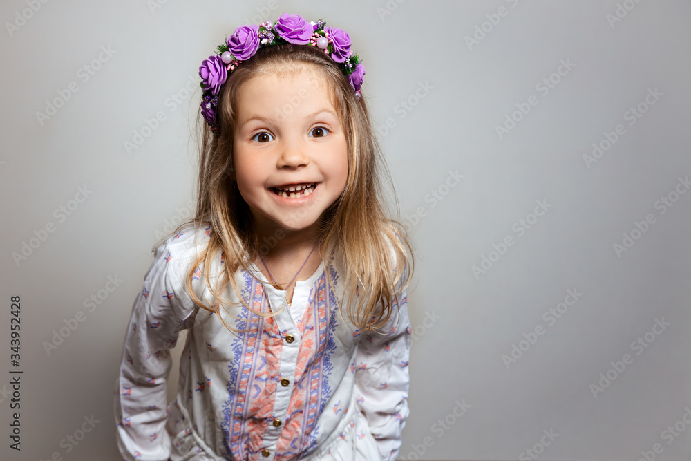 Portrait of pretty little girl smiling in white dress and purple wreath on gray isolated background. Happiness emotion concept Portrait of pretty little girl smiling in white dress and purple wreath on gray isolated background. Happiness emotion concept