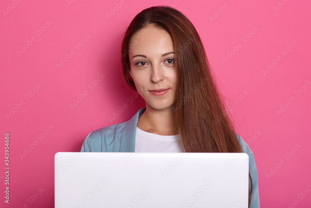 Picture of cheerful adorable girl with beautiful hair looking directly at camera, sitting in front of opened white lap top, winsome girl posing isolated over pink studio background. People concept.