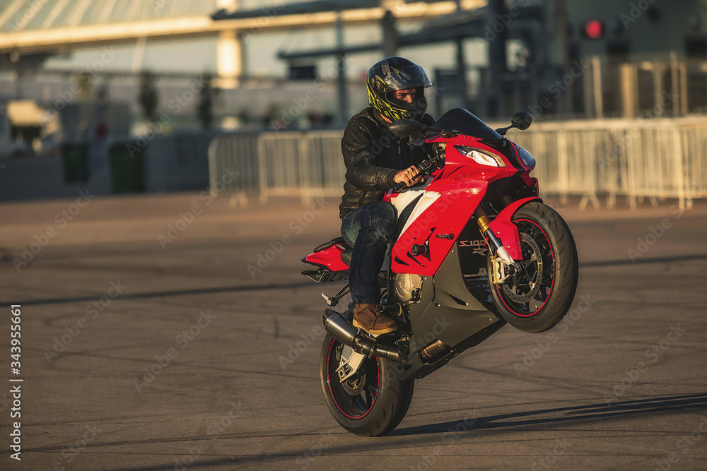 Stunt rider in helmet and black leather jacket riding red sports motorbike on one wheel