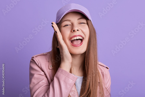 Indoor shot of smiling girl talking to somebody with happy facial expression and smile, keeping palm near mouth, wearing pink leather jacket and cap, posing isolated over lilac studio background.