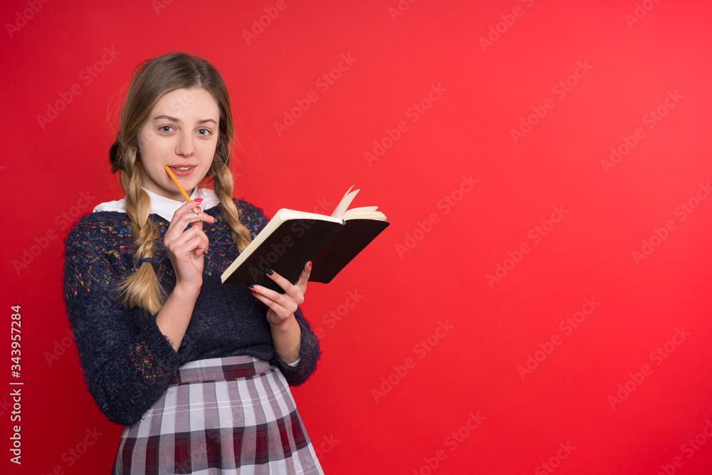 schoolgirl teen with two pigtails in a jumper and checkered skirt with a book in hand on a red background schoolgirl teen with two pigtails in a jumper and checkered skirt with a book in hand on a red background
