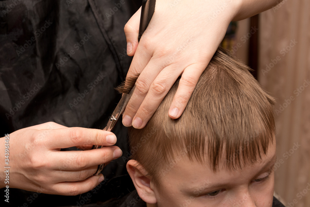 Fototapeta premium a mother-hairdresser in a black apron with a comb and scissors in her hands at home does her hair for her son, who is sitting on a chair and wrapped in a black blanket. quarantine self-isolation