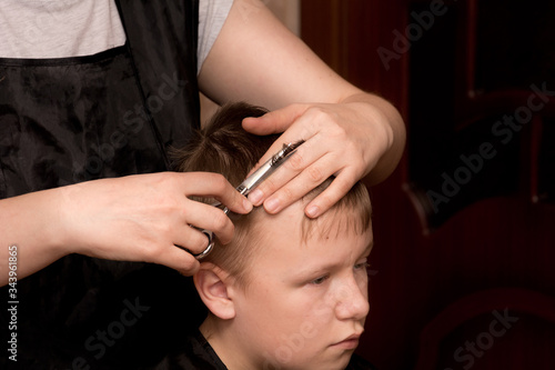 a mother-hairdresser in a black apron with a comb and scissors in her hands at home does her hair for her son, who is sitting on a chair and wrapped in a black blanket. quarantine self-isolation