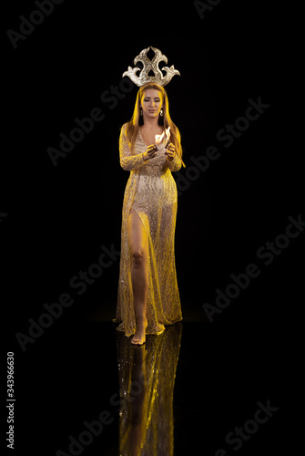 Young woman dancer in a gold dress and with a golden crown on her head holds fire in her hands on a black background
