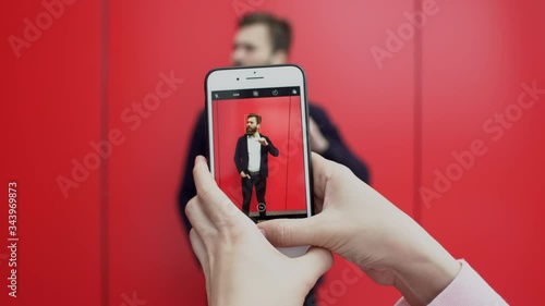 Person is taking pictures of posing near red wall brunette bearded man on the phone. Young funny guy is posing for photographer with smartphone against red background.