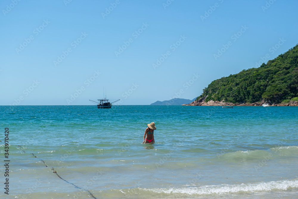 View of a beautiful beach in Brazil with coast with beautiful trees, turquoise sea and clear sky. Unrecognizable woman in bikini with hat coming out of the sea. Space for text and advertisement.