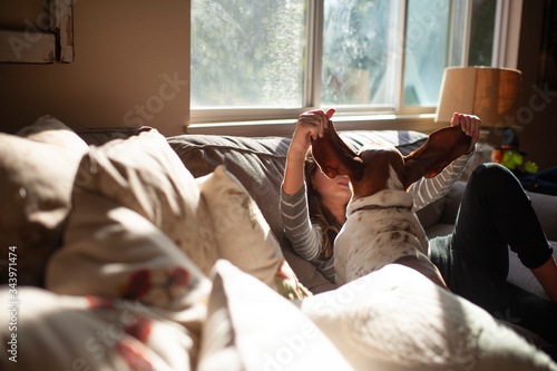 Girl sitting on the couch with basset hound dog holding out his ears