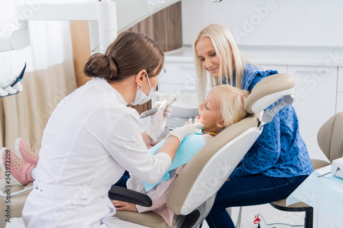 A child with a mother at a dentist s appointment. The girl lies in the chair with her mouth open  with her mother behind. The doctor is doing her job