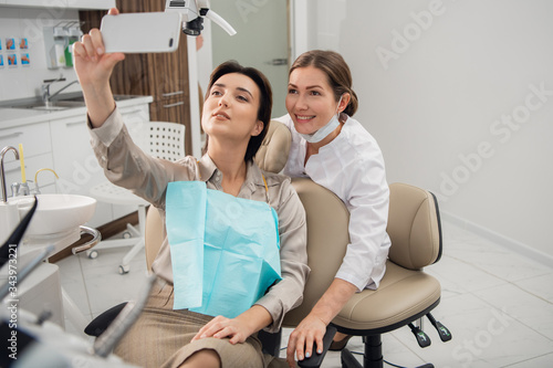 Young beautiful medical doctor  dentist  hygienist or orthodontist and her patient taking selfie with a smartphone after a successful treatment