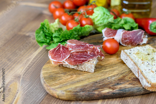 White bread and slices of smoked pork ham on the wooden kitchen board. Vegetables, green and olive oil on the kitchen table. Healthy tasty food with minerals and vitamins.