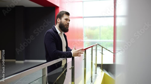 Impatient bearded man is checking the time on the watch and waiting for someone inside contemporary office building. Brunette man in black jacket and white shirt is looking forward for someone