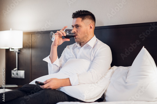 A handsome young businessman drinking red wine in bed and watching TV