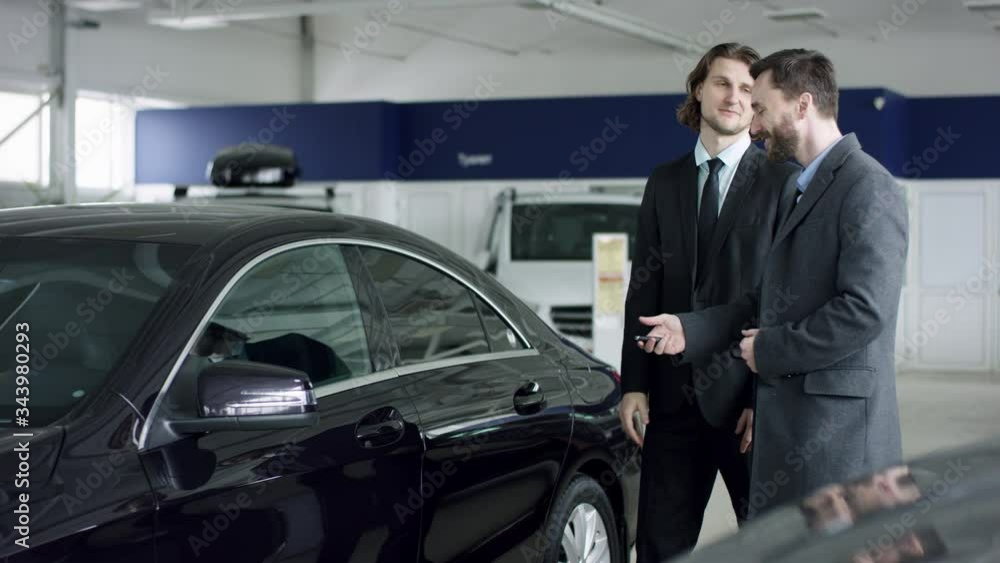 Auto dealership customer opens car door. An elegant man is choosing new automobile. A salesman invites a buyer to get in the car in showroom Auto dealership customer opens car door. An elegant man is choosing new automobile. A salesman invites a buyer to get in the car in showroom