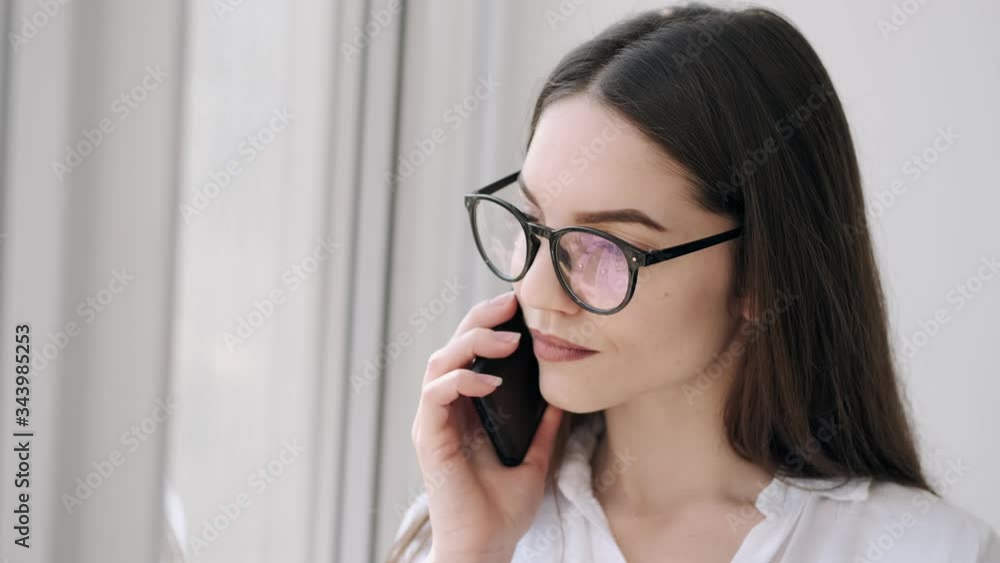A beautiful girl in glasses is talking on the phone. She is standing near the window an smiling while the communication. A beautiful girl in glasses is talking on the phone. She is standing near the window an smiling while the communication.