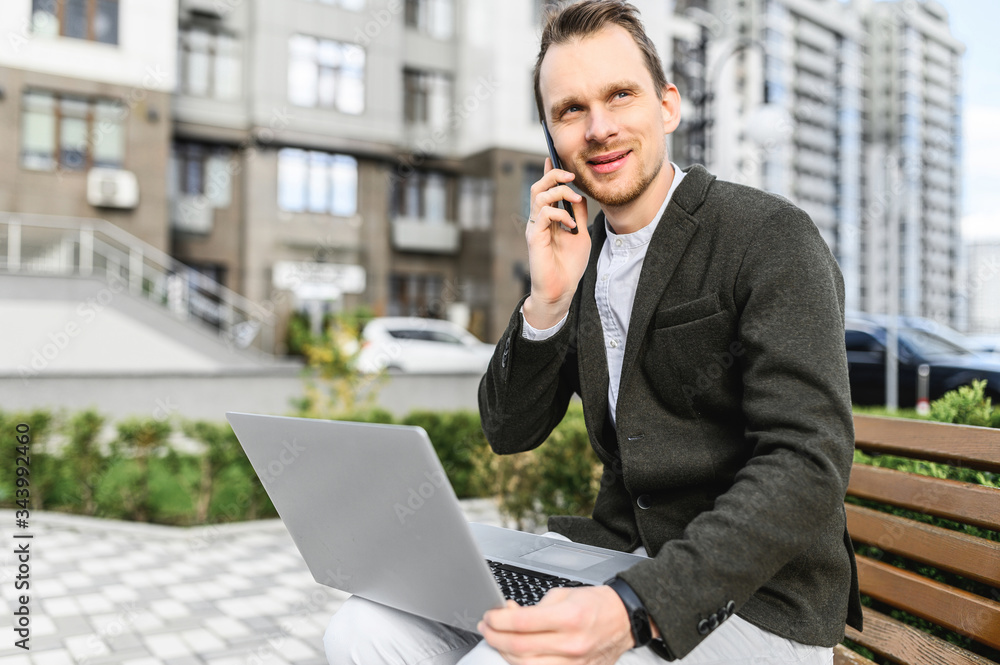 A young businessman in smart casual uses laptop for work outdoors. He sits on the bench among cityscape, looking at laptop screen and talking on the phone A young businessman in smart casual uses laptop for work outdoors. He sits on the bench among cityscape, looking at laptop screen and talking on the phone