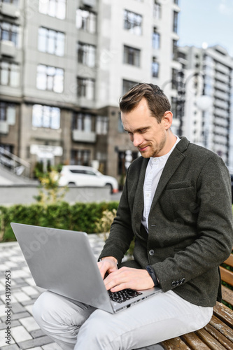 A young attractive guy uses laptop for remote work outdoors. He sits on the bench among cityscape, looking in the screen with smile and typing. Vertical photo