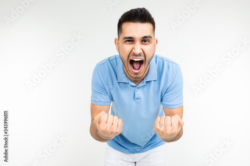handsome Caucasian in a blue T-shirt rejoices victory holding his fists on a white studio background