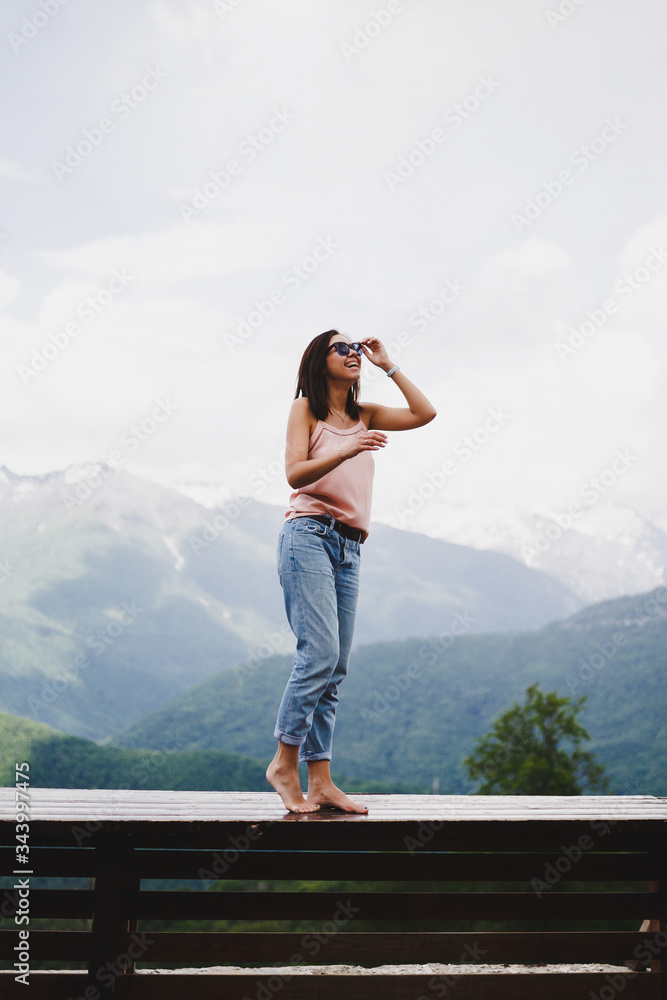 Naklejka premium A young beautiful girl in the mountains enjoys nature. The girl puts on sunglasses, smiles and enjoys a Sunny summer day.