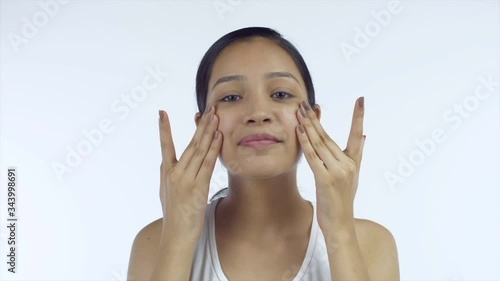 Beautiful girl rubbing moisturizer on her face using her hands - Skincare Concept. Smiling Indian female with no makeup applying night face cream on her cheeks before sleeping white background photo