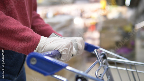 Woman in a supermarket rolls a grocery cart in a supermarket close-up of white rubber gloves. Protects against coronavirus. Purchase products for self-isolation and quarantine.