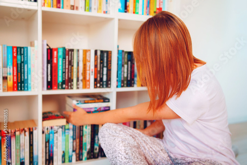 Portrait of young adult caucasian woman female girl in front of the bookshelf at home during the weekend or holiday picking the book to read from the shelf wearing eyeglasses back view