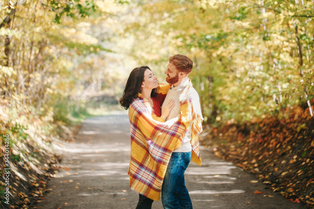 Fototapeta premium Beautiful couple man woman in love. Smiling laughing boyfriend and girlfriend wrapped in yellow blanket hugging in a park on autumn fall day. Togetherness and happiness. Authentic real people.